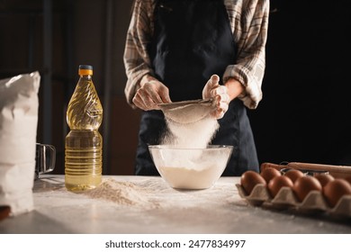 woman sifting flour through sieve into transparent bowl while baking at home. Flour dust visible in the air, dynamic scene. Baking ingredients such as eggs, flour and butter are placed on the table. - Powered by Shutterstock