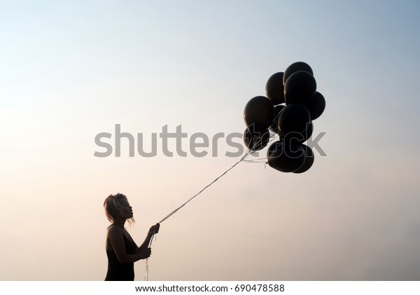 Woman in\
silhouette looking at black balloons about to let go set against a\
stark clean background and sky at\
sunset
