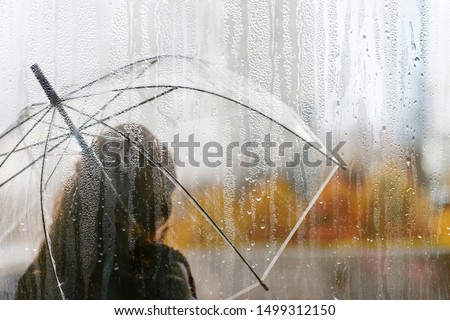 A woman silhouette with transparent umbrella through wet window with drops of rain. Autumn 