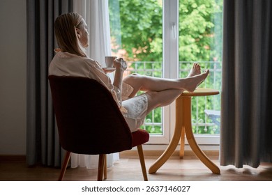 A woman in a silk robe enjoys a peaceful morning, sipping coffee while seated by a large window. Her legs rest on a small wooden table as she gazes outside at the lush greenery. 
