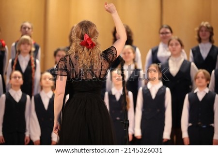 Woman singer blonde in black dress with raised hand leads the choir standing in front of a group of children students of a music school back view of the leader