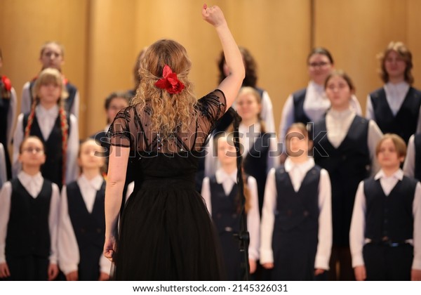 Woman singer blonde in black\
dress with raised hand leads the choir standing in front of a group\
of children students of a music school back view of the\
leader