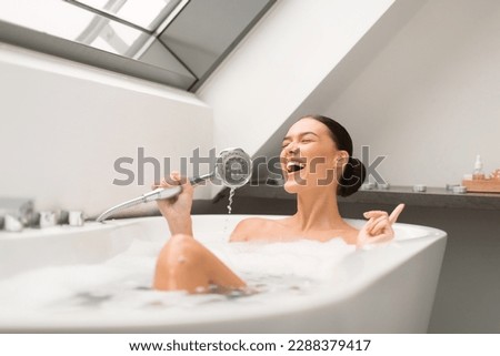Woman Singing Taking Bath With Foam, Having Fun Holding Shower Head Like Microphone Relaxing In Modern Bathroom Indoor. Cheerful Female Enjoying Bathing With Eyes Closed At Home