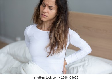 Woman sit in bed awakening get out from bed feeling lower back pain frown touch or massaging lumbus to relieve ache caused by incorrect sleeping posture, unsupportive mattress, disease symptom concept - Shutterstock ID 1569483655