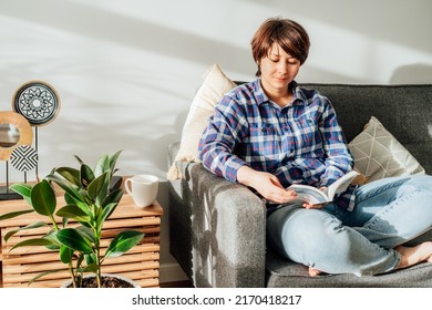 Woman sit on the sofa couch and relax while reading a book at home in cozy modern interior living room with green plants. Offline activity lifestyle. Reading hobby. Leisure indoors entertainment