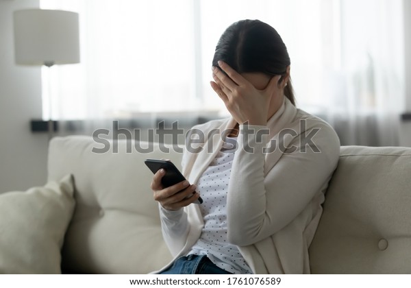 Woman sit on sofa holding smartphone cover face\
with hand feels scared humiliated suffering from cyberbullying\
being on-line abused by stalker. Bad news, life troubles, break up\
with boyfriend concept