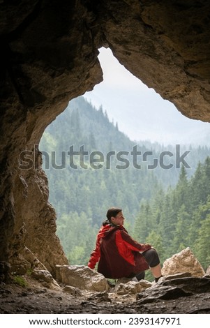 a woman sits by a cave and looks at the mountain landscape, view from inside the cave in summer