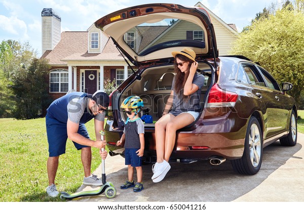 Woman\
sits in cars trunk and smiling.Men and toddler boy standing near\
vehicle, holding kickboard.Happy family time.Mother,father,son near\
house in suburban neighborhood.Warm\
weather,summer.