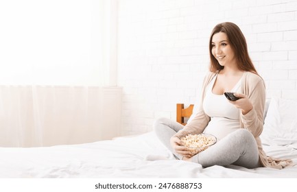 A woman sits comfortably on a bed holding a remote and eating popcorn while watching television. She is dressed in casual lounge wear, enjoying a relaxing morning in a brightly lit bedroom - Powered by Shutterstock