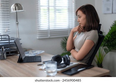 A woman sits at a desk with a laptop and a keyboard. She is wearing a gray shirt and has her hands clasped behind her back. The room has a window with blinds and a potted plant - Powered by Shutterstock