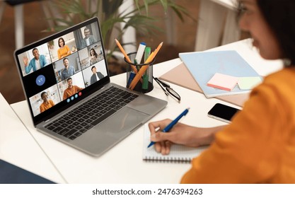 A woman sits at a desk with a laptop open, displaying a video conference meeting with several participants. She is taking notes in a notebook with a blue pen, her focus on the screen. - Powered by Shutterstock