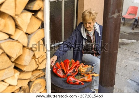 A woman sits in front of the house, roasting peppers for the delicacy ajvar in the yard on an autumn day. She roasts the peppers on the burner of a wood fired stove