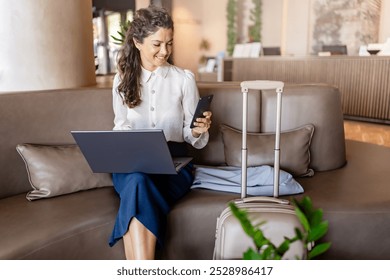 A woman sits in a hotel lobby using a laptop and smartphone while her suitcase stands nearby. She appears focused on her work in a comfortable, modern lounge area. - Powered by Shutterstock