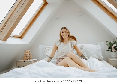 A woman sits on a bed by a skylight, smiling with comfort on a sunny day