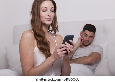 woman sits on the bed and writes a message on her smart phone. her partner makes a jealous and disappointed and frustrated expression. 