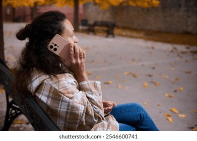 A woman sits on a bench in an autumn park, talking on her smartphone. The surroundings are filled with falling leaves and warm colors, creating a peaceful and relaxing atmosphere. - Powered by Shutterstock