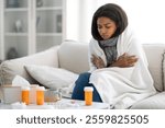 A woman sits on a couch wrapped in a blanket, looking cold and unwell. Medication bottles and tissues are on the table nearby, highlighting her struggle with illness during a cold day, copy space