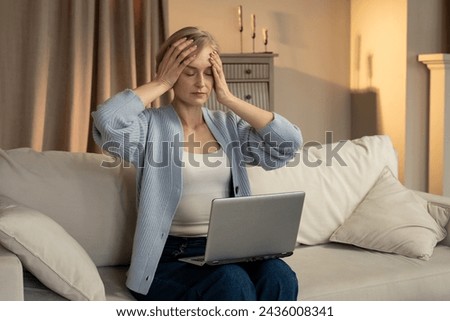 A woman sits on a cream-colored sofa, her expression one of frustration or exhaustion. She holds her hand over her face in a gesture of dismay, as a laptop rests on her lap, suggesting difficulty or