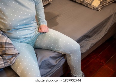 A woman sits on the edge of the bed in wet pyjamas. Wet stain on grey sheets. Incontinence. Nocturnal enuresis-inability to control the act of urination. Concept.