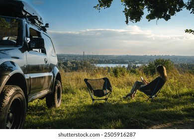 A woman sits on a folding, portable chair and enjoys the moment of life. Nature, on the top, river, sunset. Tourism and active lifestyle. Equipment for tourism. Off road vehicle - Powered by Shutterstock