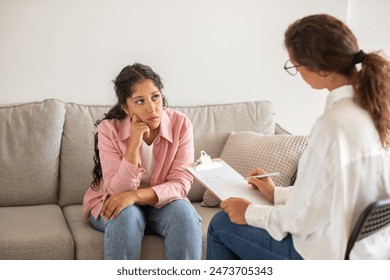 A woman sits on a gray couch in a white room while a therapist listens and takes notes. The woman looks thoughtful and concerned while the therapist appears attentive. - Powered by Shutterstock