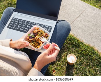 Woman sits on park bench with laptop and take away lunch box, cardboard cup of coffee. Healthy bowl with vegetables. Casual clothes, urban lifestyle of millennials. Healthy nutrition. 