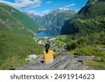 A woman sits on a rocky ledge overlooking a breathtaking Norwegian fjord, taking in the beauty of the green mountains and blue waters. Geiranger Fjord Norway