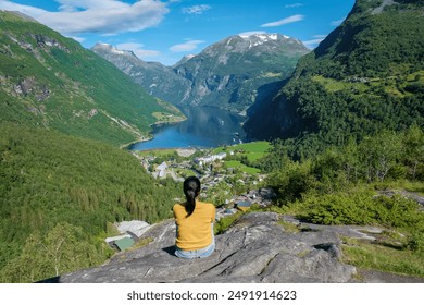 A woman sits on a rocky ledge overlooking a breathtaking Norwegian fjord, taking in the beauty of the green mountains and blue waters. Geiranger Fjord Norway - Powered by Shutterstock