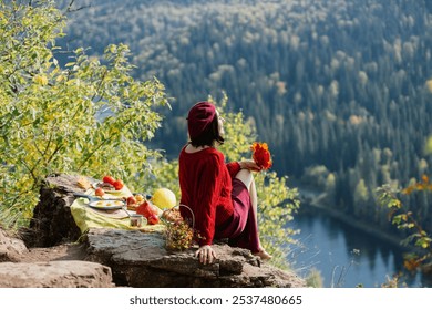 A woman sits on a rocky ledge, admiring the picturesque view of a calm lake framed by vibrant autumn trees. A picnic spread rests on the ground nearby, enhancing the peaceful atmosphere. - Powered by Shutterstock