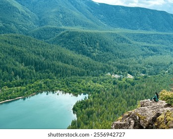 A woman sits on a rocky ridge overlooking a tranquil lake, dense forests, and distant mountains under a moody sky in Washington state - Powered by Shutterstock