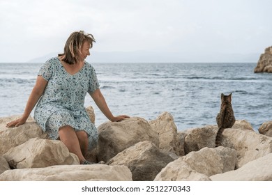 Woman sits on  rocky seashore and looks at  street cat. Joyful life after 50. Vacation, travel.  - Powered by Shutterstock