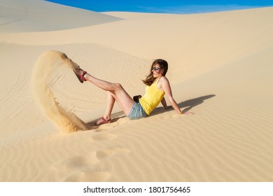Woman sits on the sand in the desert, wearing a T-shirt and shorts. Playing with sand.