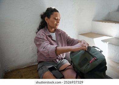 A woman sits on stairs packing a backpack, dressed casually, with a thoughtful expression, in a cozy and relaxed setting. - Powered by Shutterstock