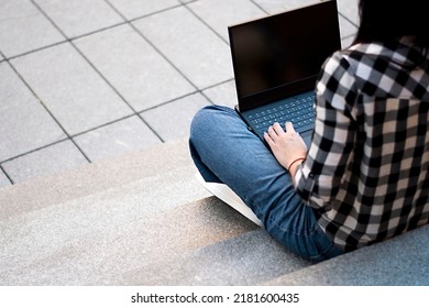 A Woman Sits On The Steps In A Black And White Checkered Shirt Outside And Holds A Black Laptop On Her Lap.