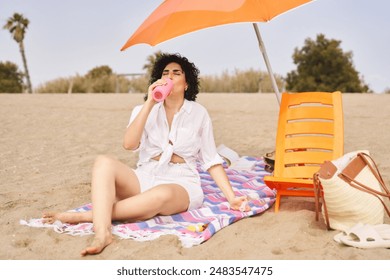 A woman sits on a towel on the beach, drinking water from a pink bottle, hydrating against the heat - Powered by Shutterstock