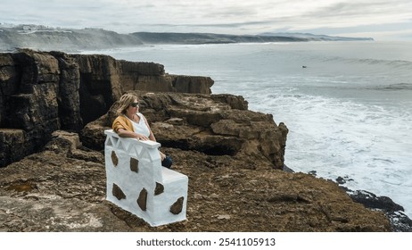 A woman sits on a white bench on the rocky shore of the Atlantic Ocean in Ericeira, Portugal, looking out into the vast waters - Powered by Shutterstock