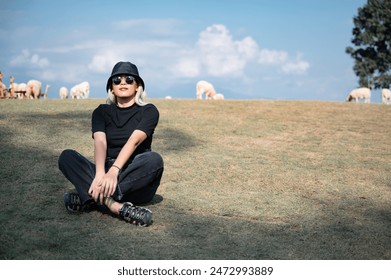 A woman sits outdoors on the grass. with sheep grazing in the background The person is wearing sunglasses. Black bucket hat, black t-shirt, and dark pants. And they looked relaxed and content. - Powered by Shutterstock