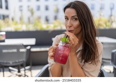 woman sits outdoors, sipping a refreshing pink drink from a mason jar with a black straw and mint garnish. The background features a sunny outdoor cafe with plants and blurred buildings. - Powered by Shutterstock