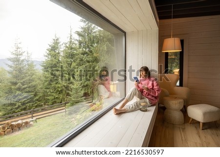 Woman sits with phone on window sill and enjoys scenic view on mountains while resting in wooden house on nature. Recreation and escaping to nature concept