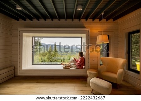 Woman sits with phone on window sill and enjoys scenic view on mountains while resting in wooden house on nature. Wide interior view of cozy room
