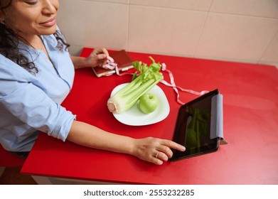 A woman sits at a red table, interacting with a tablet. Fresh vegetables and an apple are on the table, symbolizing a focus on healthy living and diet planning with digital tools. - Powered by Shutterstock