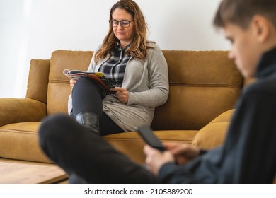 A Woman Sits In The Waiting Room And Reads A Magazine. A Young Man Looks At His Phone In The Blurred Foreground