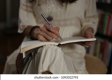 Woman sits in a white dress with a leather journal open on her lap, writing with a pen. 