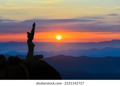 A woman sits in a yoga pose on a mountain peak, arms raised, silhouetted against a vibrant sunrise over misty hills. - Powered by Shutterstock