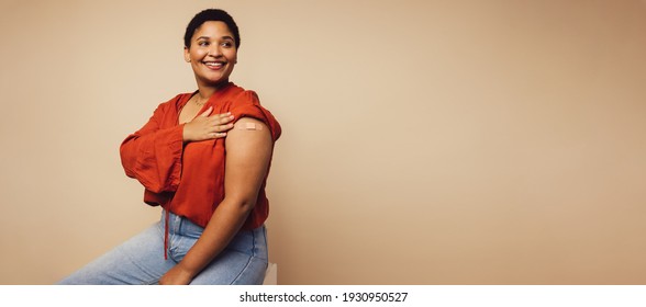 Woman sitting against brown background showing her arm with bandage. Woman received a vaccine.