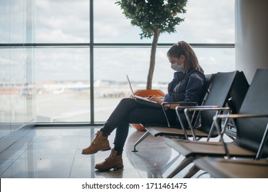 A Woman Is Sitting At The Airport With A Laptop In A Medical Mask. A Young Girl Is Waiting For Departure At The Gate, Working Online. Social Responsibility, Virus Protection.
