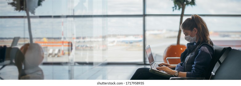 A Woman Is Sitting At The Airport With A Laptop In A Medical Mask. A Young Girl Is Waiting For Departure At The Gate, Working Online. Social Responsibility, Virus Protection.