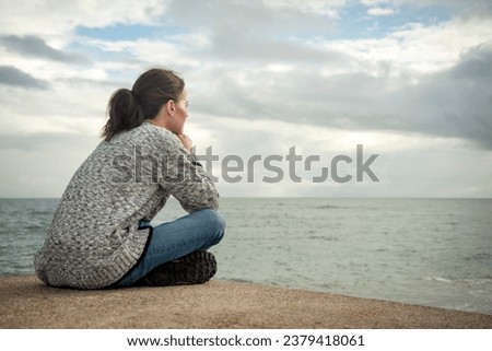 Woman sitting alone looking out to sea deep in thought, rear view, Autumn.