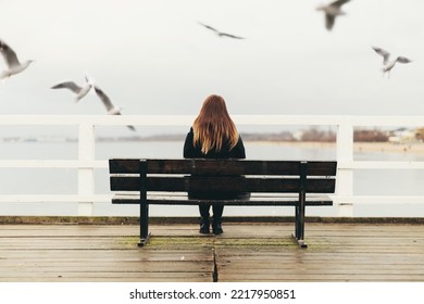 Woman sitting alone on bench by the sea. Gulls flying