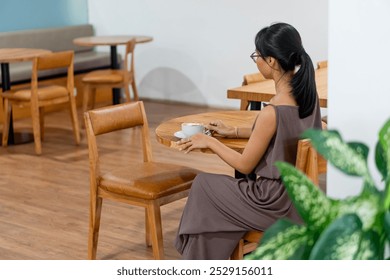 A woman sitting alone at a wooden table in a cozy cafe, wearing a brown dress and glasses. She holds a cup of coffee, looking contemplative. The cafe has a minimalist eds - Powered by Shutterstock